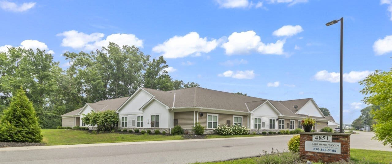 Front view of Lakeshore Woods Seniors hospice care facility surrounded by greenery and a clear blue sky, offering a peaceful and welcoming environment for residents.