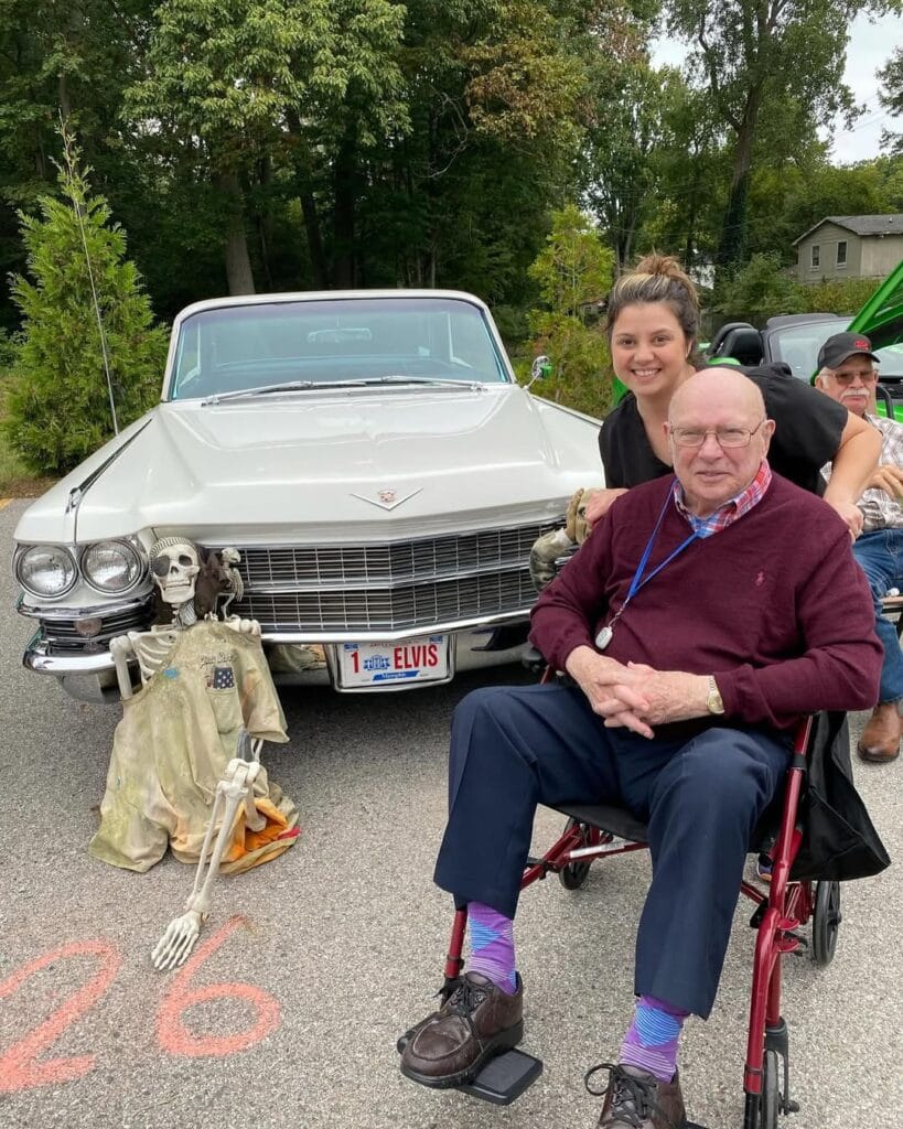 A Lakeshore Woods resident in a wheelchair and a staff member smiling beside a vintage white Cadillac at an outdoor classic car show.