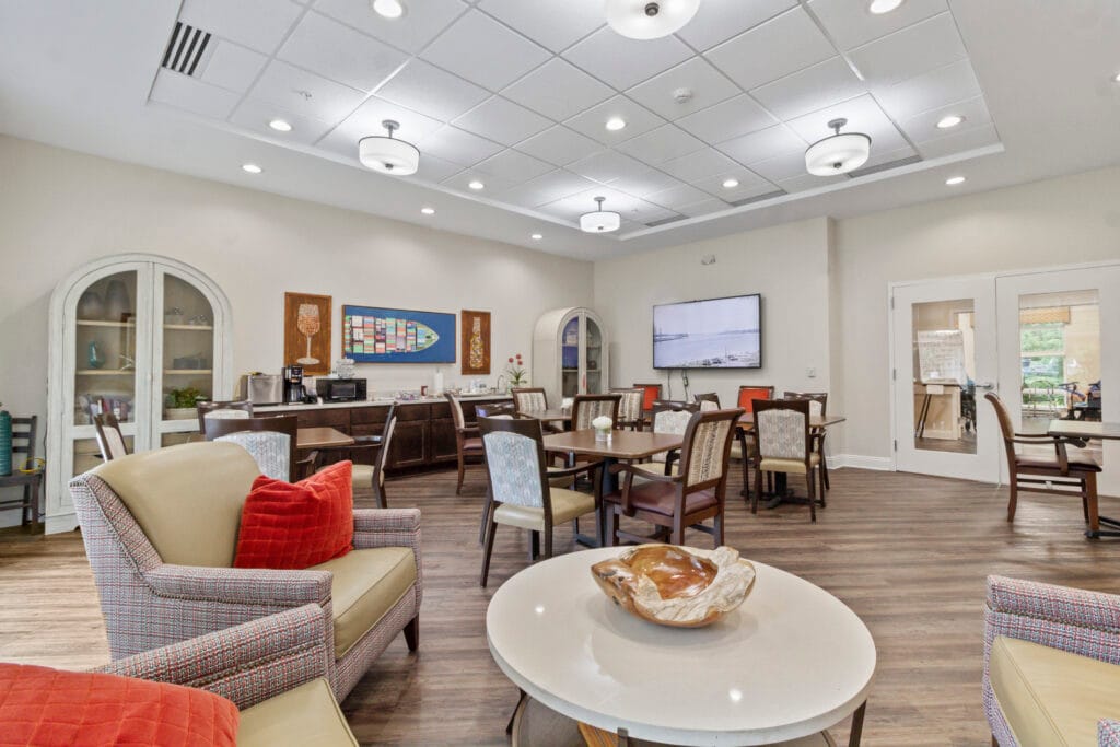 Community lounge at Lakeshore Woods with armchairs, dining tables and a wall-mounted television.