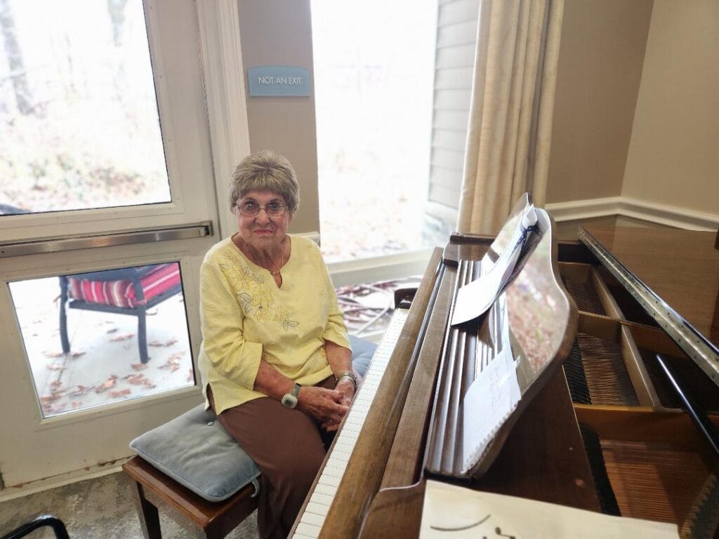 A Lakeshore Woods resident seated at an upright piano beside a sunlit window.